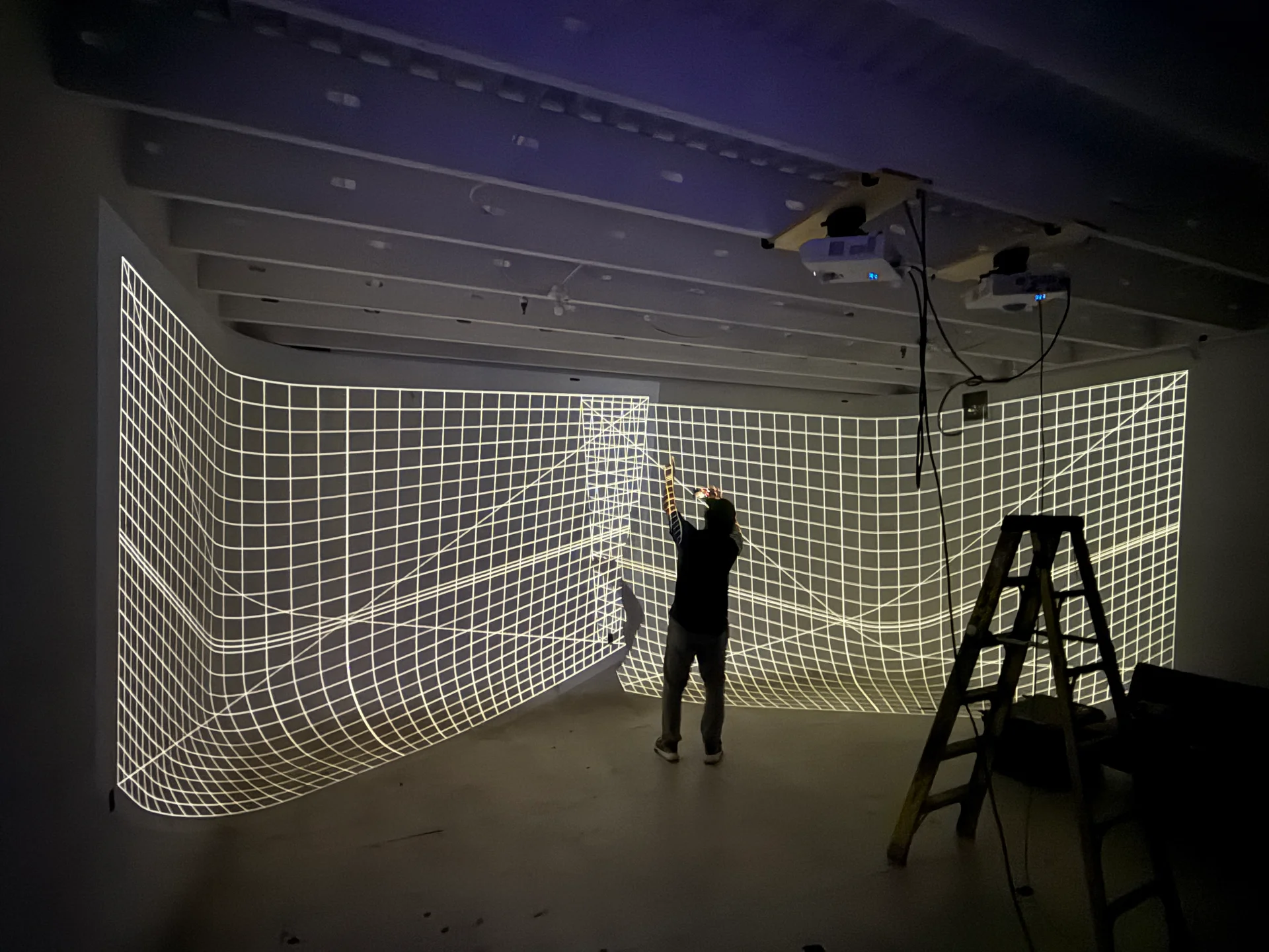 A person reaches toward a projected calibration grid during installation setup, with ceiling-mounted projectors and a ladder nearby.